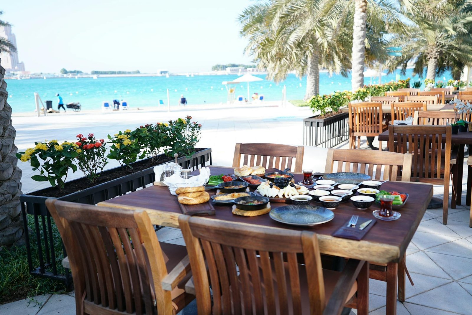 Elegant dining setup by a turquoise sea, surrounded by lush palm trees and vibrant flowers.