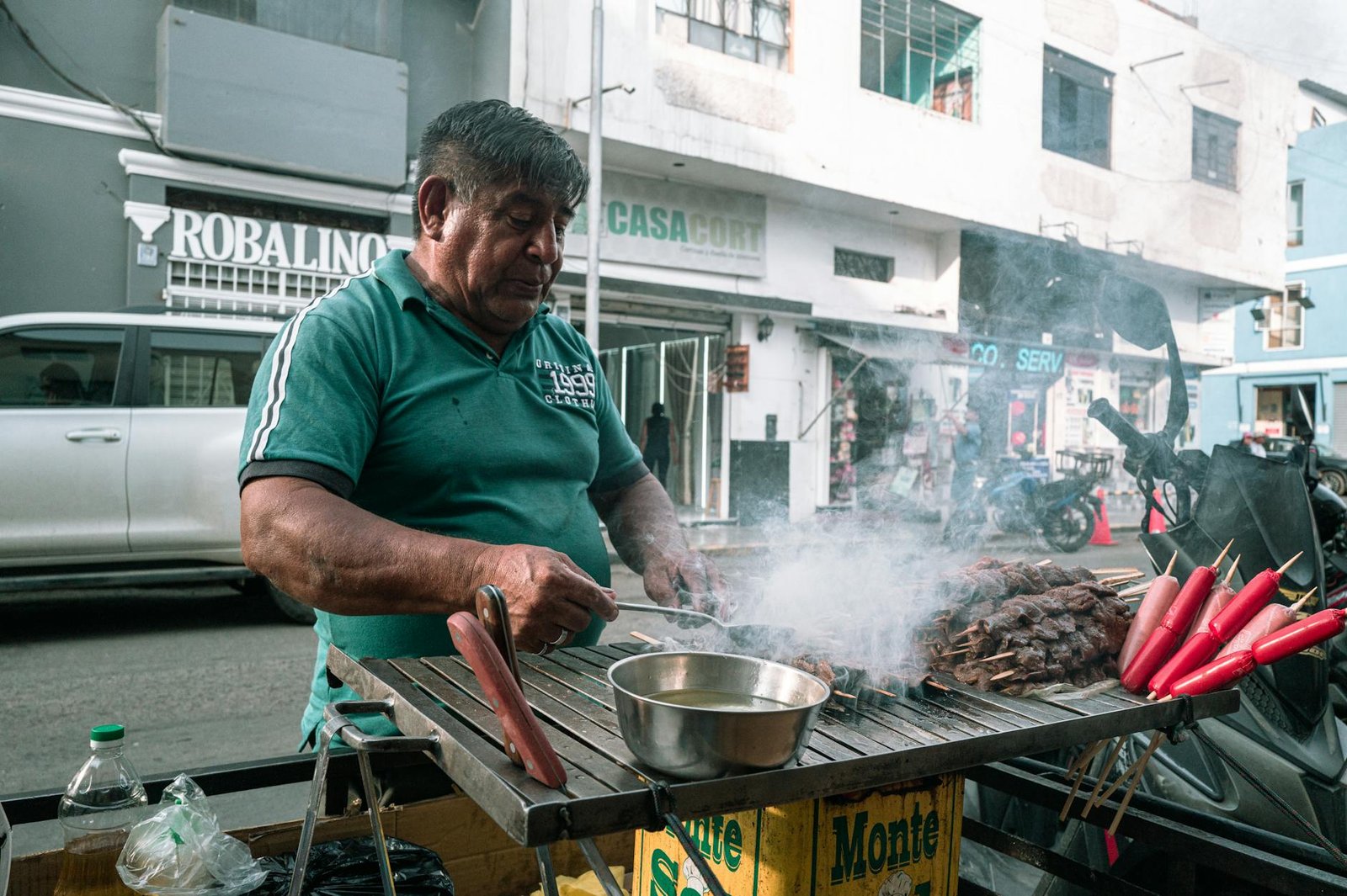 A street vendor grills skewers outdoors in an urban Peruvian setting with smoke rising.