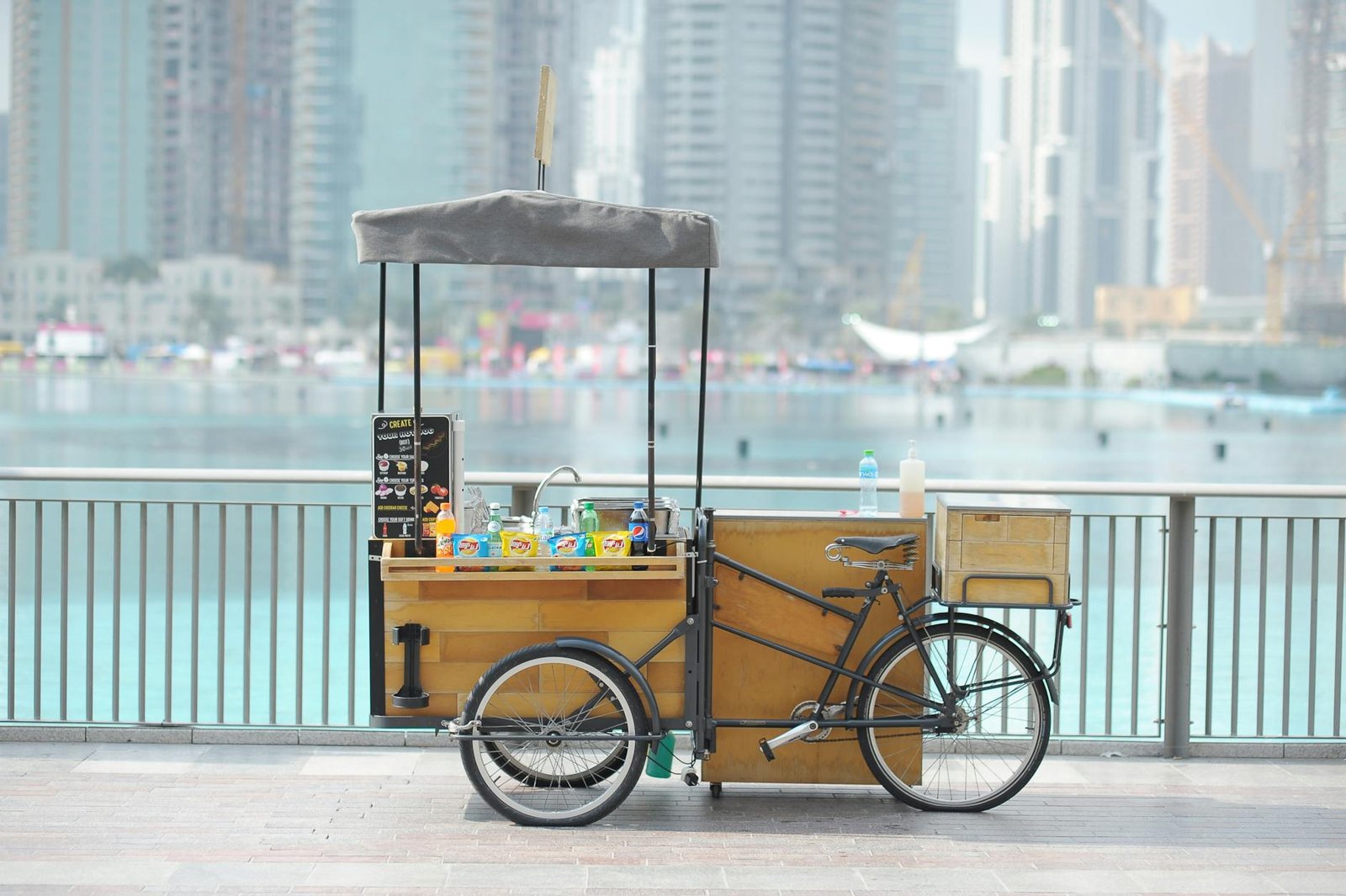 A bicycle vendor cart selling drinks by the waterfront with Dubai skyline in the background.