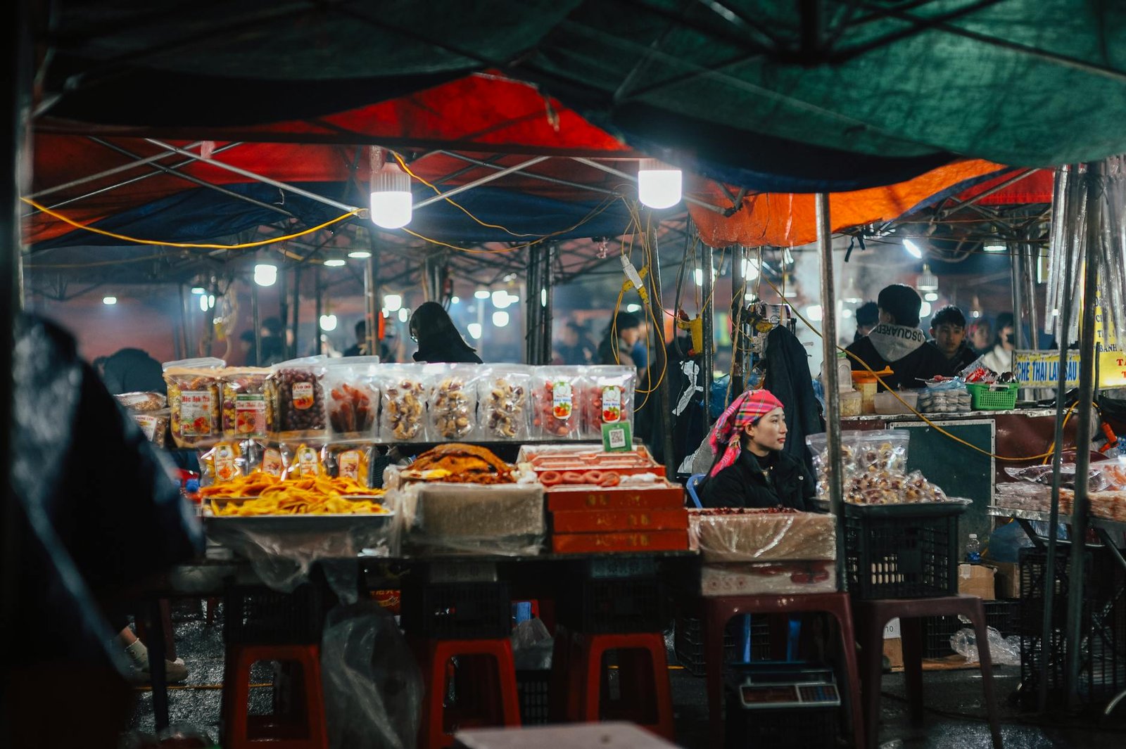 A bustling night market scene featuring vendors with various food items, illuminated by colorful lights.