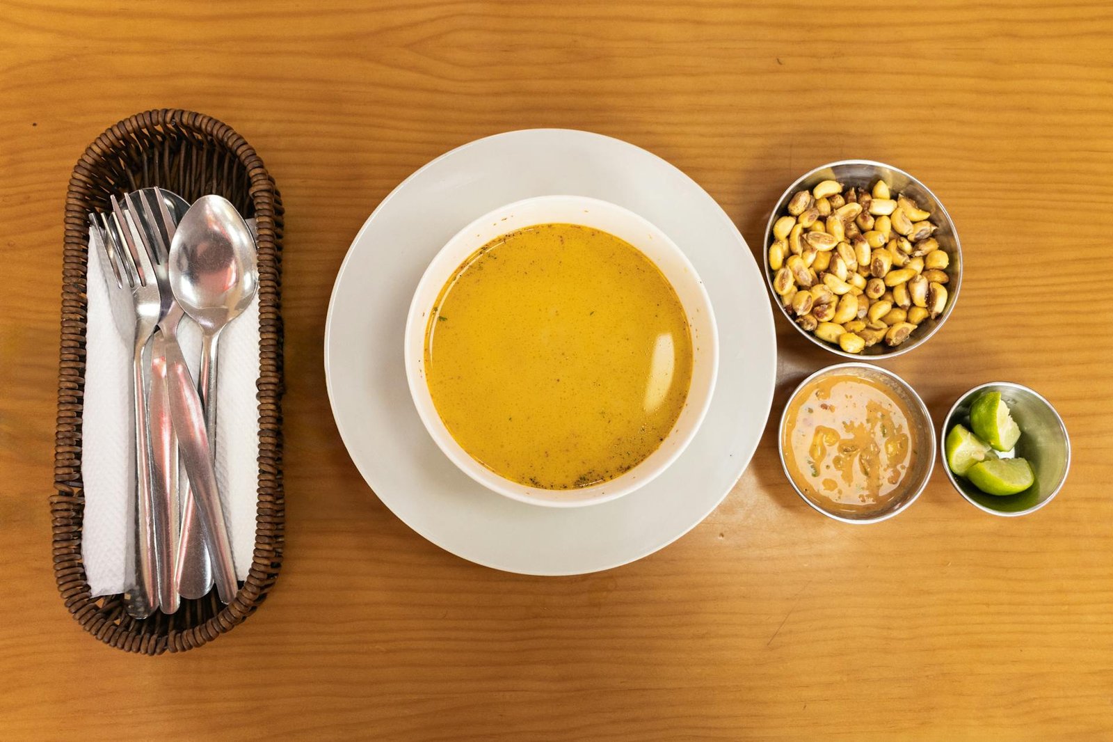 A delicious bowl of Peruvian soup with sides of corn, sauce, and lime on a wooden table.