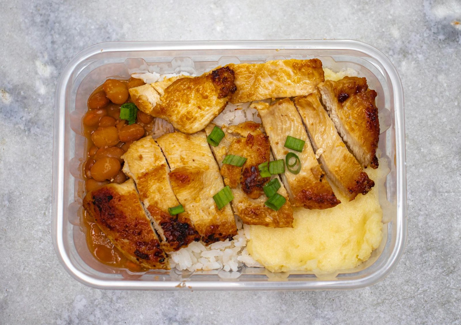 Grilled chicken with rice, beans, and mashed potatoes in a takeout container.