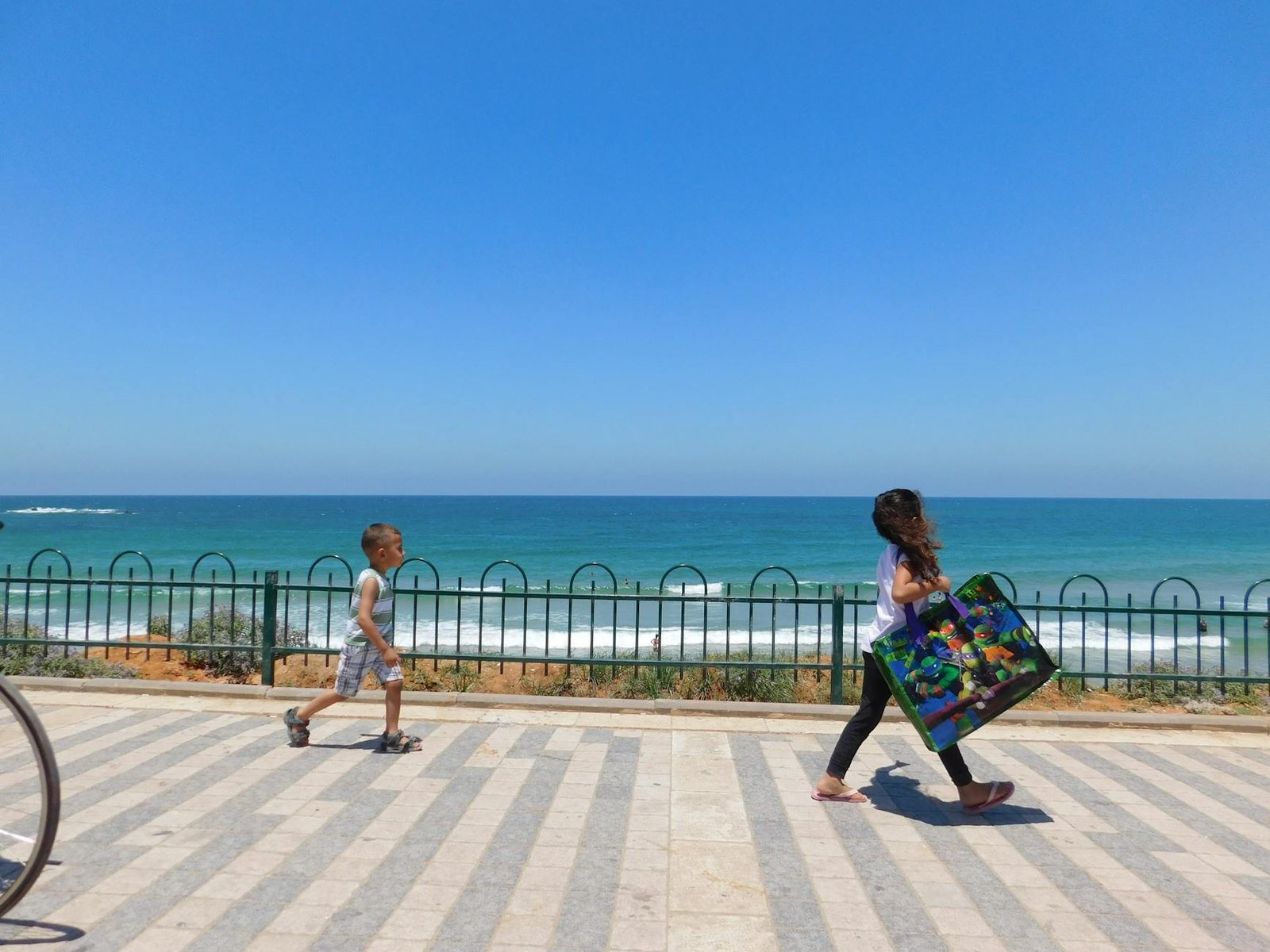 Two people walk along the seaside promenade in Tel Aviv, Israel, under a clear blue sky.
